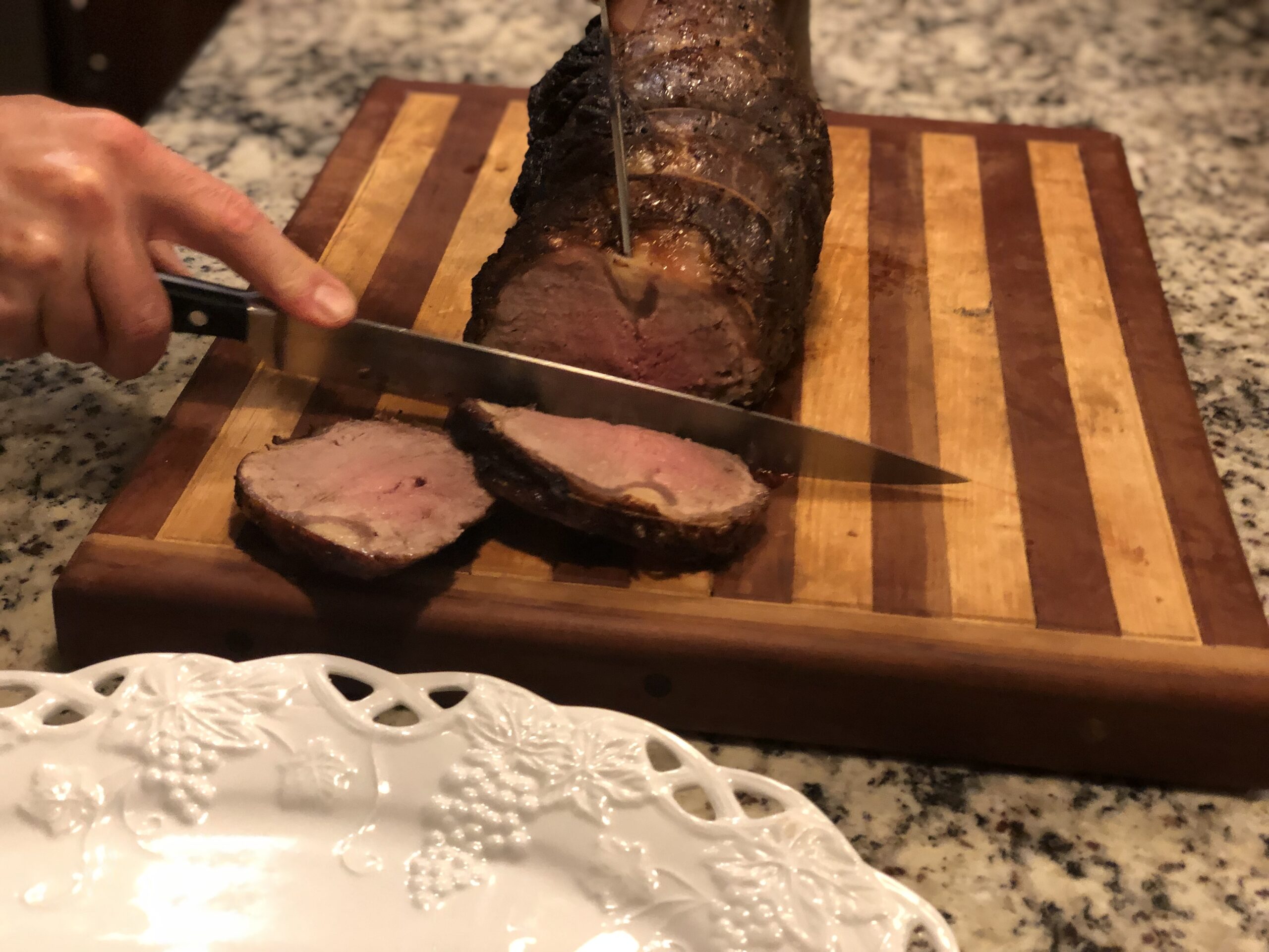 A person slices a perfectly cooked roast beef on a wooden cutting board, showcasing the tender pink interior, with a decorative white plate in the foreground. The scene highlights a culinary moment in a home kitchen.