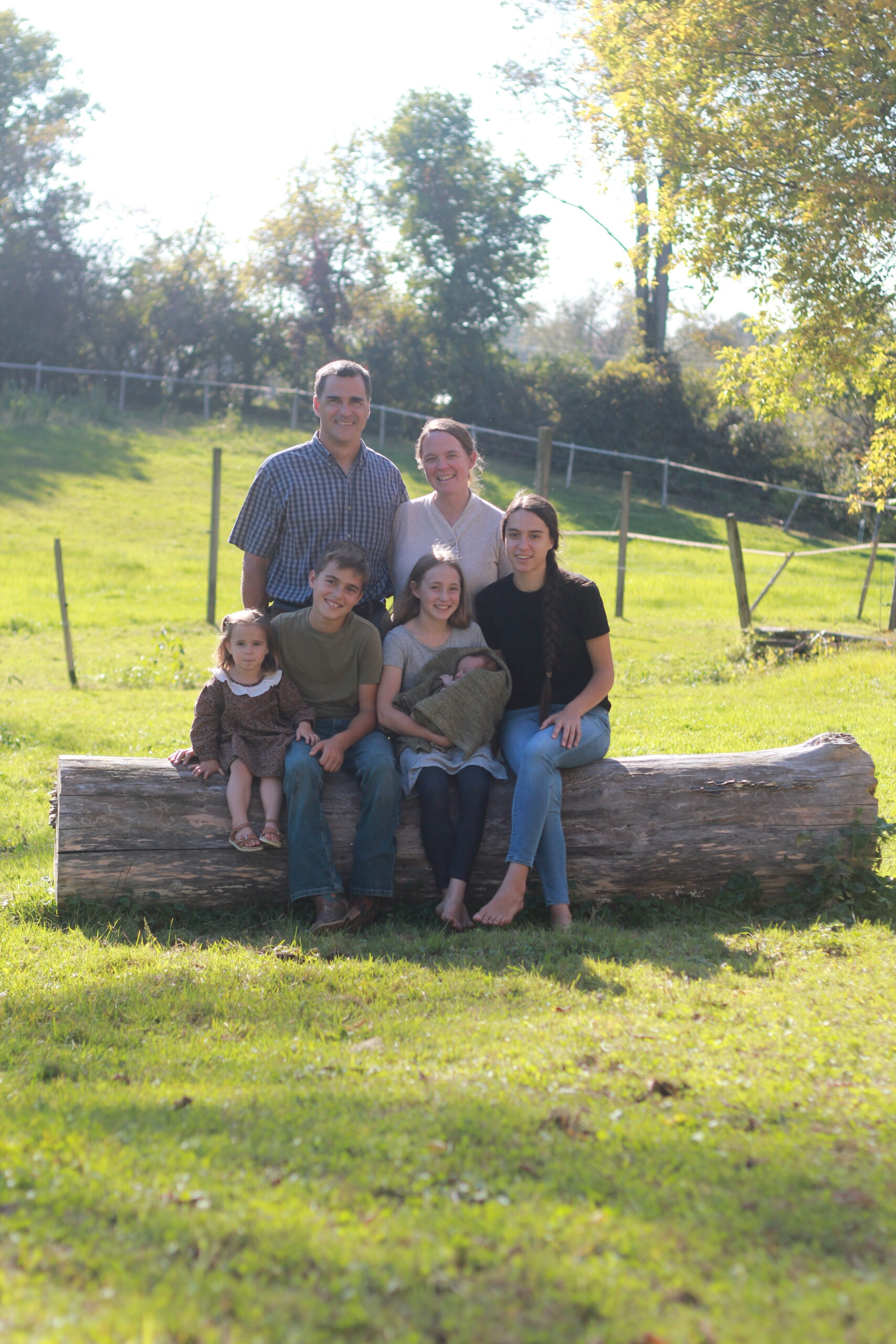 Family portrait sitting on a log in a sunny green field, featuring a smiling family of six, including two adults and four children, with a peaceful rural backdrop.