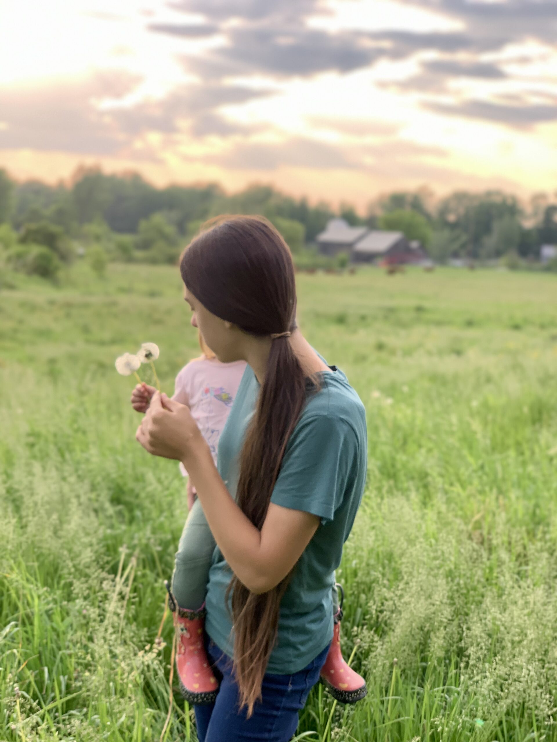 Mother carrying child in a field at sunset, both enjoying nature with dandelions, showcasing family bonding and outdoor leisure.
