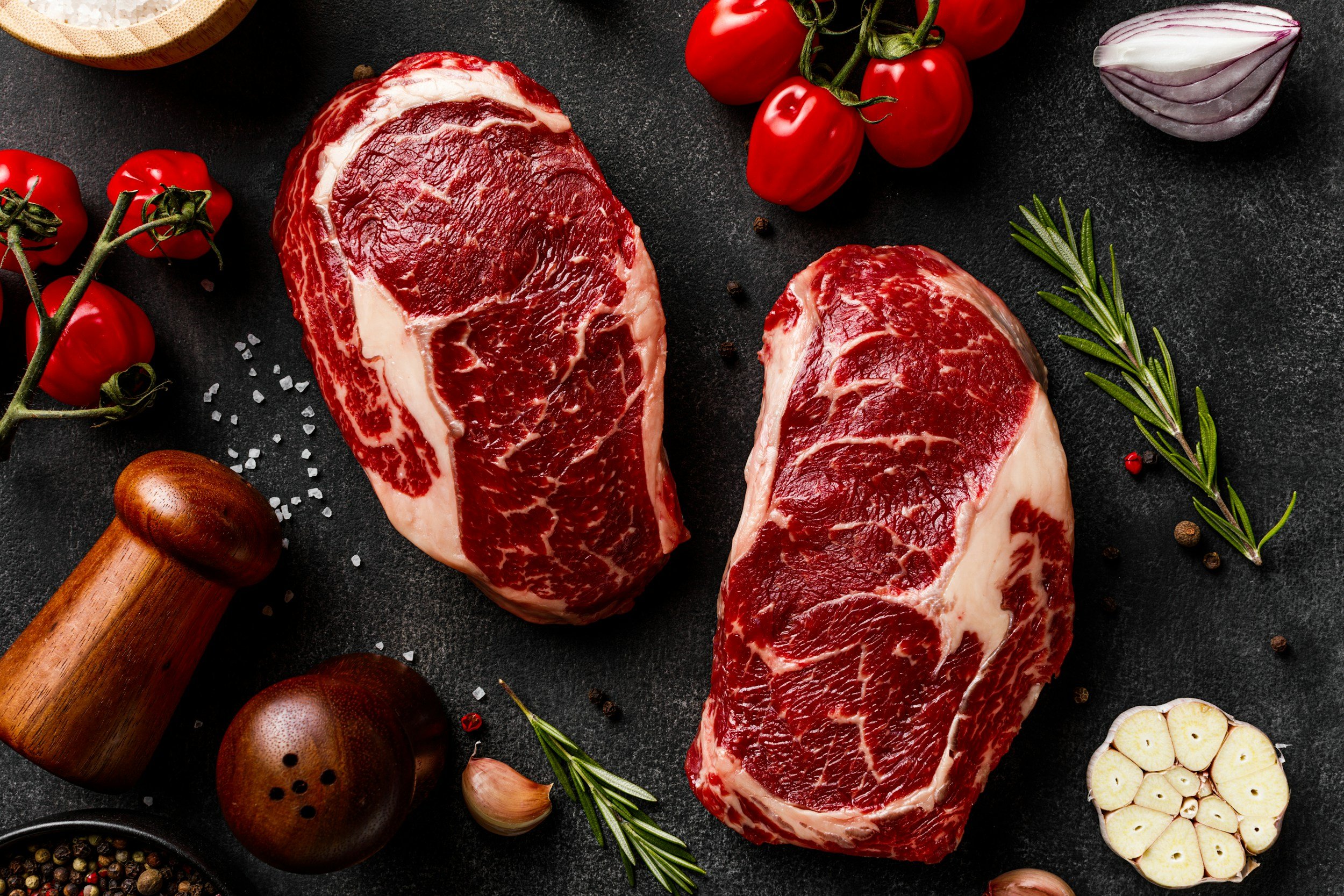 unsplash-image-j-17JLHMIpk Fresh ribeye steaks surrounded by cherry tomatoes, garlic, rosemary, and spices on a dark textured surface, ideal for gourmet cooking and grilling.