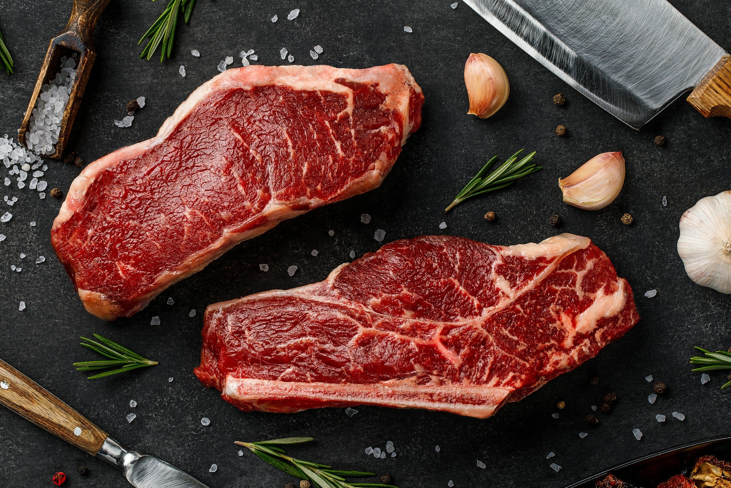 unsplash-image-sV8Cl0_3UcY Raw ribeye steaks on a dark stone surface, surrounded by garlic cloves, rosemary sprigs, coarse salt, and a chef's knife, showcasing marbling and freshness for gourmet cooking.